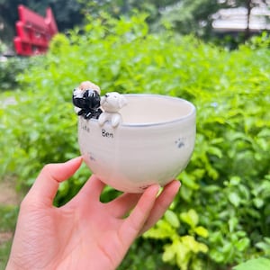 May include: A cream ceramic bowl featuring two dog figurines, one black and white, the other white, on the rim. The bowl is decorated with paw prints and the names "Luke" and "Ben".