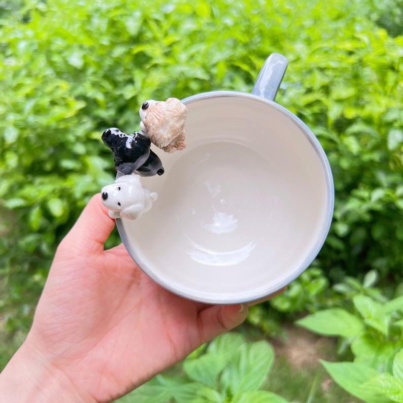 May include: A white ceramic mug with a pale blue rim and handle, decorated with three small dog figurines in white, black, and tan. The mug is held in a hand against a backdrop of green foliage.