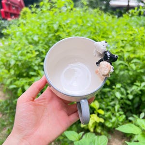 May include: A white ceramic mug with a pale blue handle and rim, decorated with three small dog figurines in black, white, and tan. The mug is held against a backdrop of green foliage.