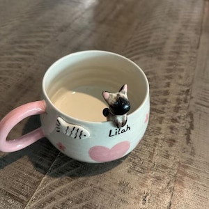 May include: White ceramic mug with a pink handle. The mug has a pink heart, a fish design, and a small cat figurine peeking over the rim. The mug is personalized with the name "Lilah".