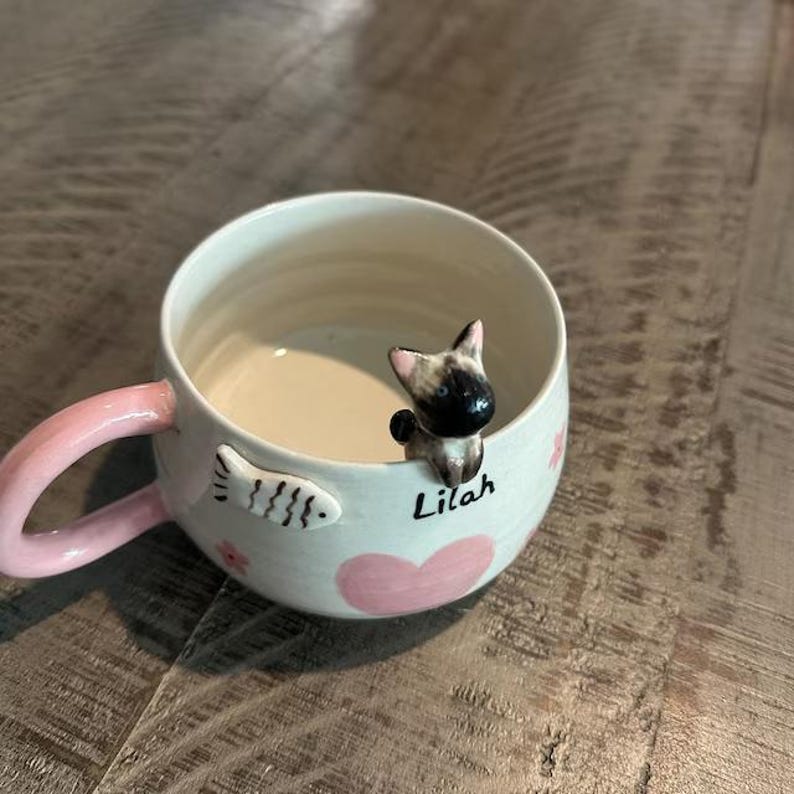 May include: White ceramic mug with a pink handle. The mug has a pink heart, a fish design, and a small cat figurine peeking over the rim. The mug is personalized with the name "Lilah".