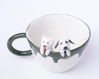 May include: A white ceramic mug with a green handle and a green rim. Two small ceramic dog figurines are perched on the rim of the mug, one white and one black and white.