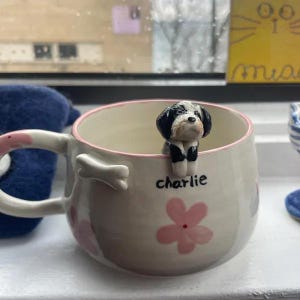May include: A white ceramic mug with pink flowers and a bone-shaped handle. A small black and white dog figurine is perched on the rim of the mug. The mug is personalized with the name "charlie".