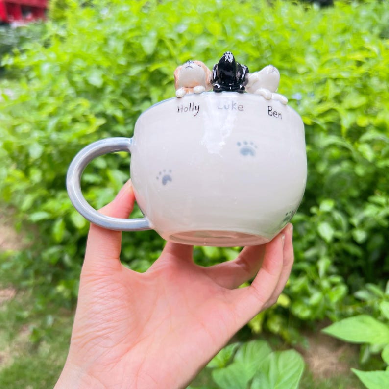 May include: A white ceramic mug with a pale blue handle and paw print details. The mug is decorated with three small dog figurines, and the names "Holly", "Luke", and "Ben" are written on the mug.