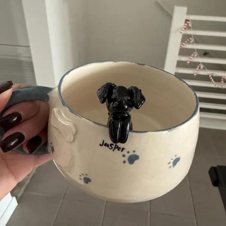 May include: A white ceramic mug with blue paw prints and the name "Jasper" written on it. A black ceramic dog figurine is sitting inside the mug.