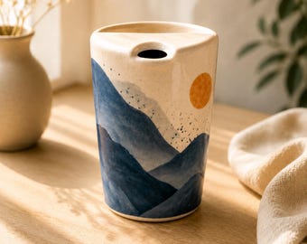 Può includere: Un vaso cilindrico in ceramica con una base color crema caratterizzato da un paesaggio dipinto di montagne blu a strati e un sole arancione brillante. Il bordo superiore presenta un design ritagliato unico e la superficie ha una finitura liscia.