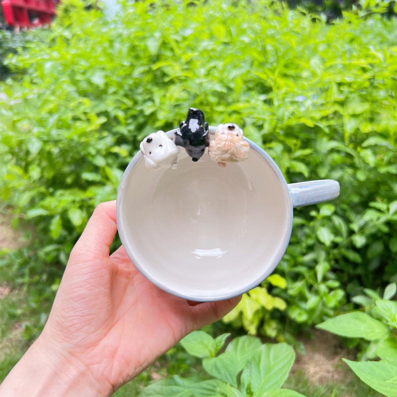 May include: A pale blue ceramic mug featuring three small dog ornaments around the rim. The dogs are white, black, and tan. The mug has a light blue handle and a white interior.