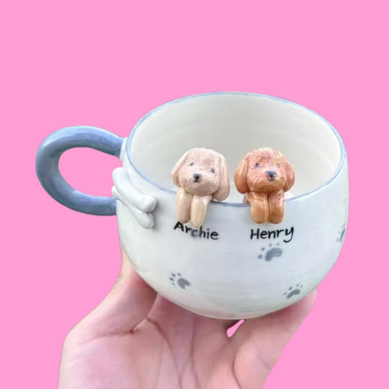 May include: A white ceramic mug with a blue handle features two small ceramic dog figurines peeking over the rim. The dog figurines are named "Archie" and "Henry" on the mug.