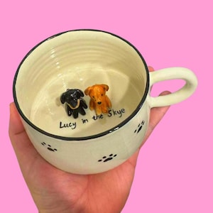 May include: A white ceramic dog bowl with black paw prints and a black rim. The bowl has two small ceramic dog figurines inside, one black and one brown. The bowl is personalized with the text "Lucy in the Skye".