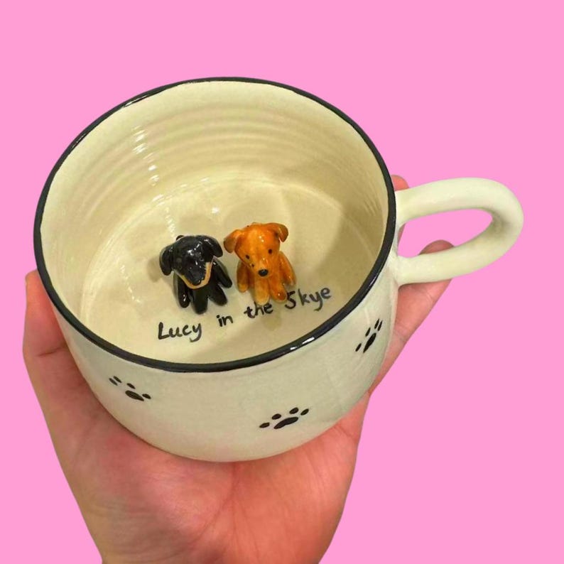 May include: A white ceramic dog bowl with black paw prints and a black rim. The bowl has two small ceramic dog figurines inside, one black and one brown. The bowl is personalized with the text "Lucy in the Skye".