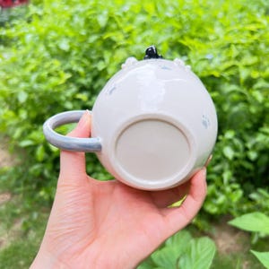May include: A white ceramic mug with a rounded shape, a pale blue handle, and paw print designs. The mug has a black cat figurine on top. The mug is held in a hand against a background of green foliage.