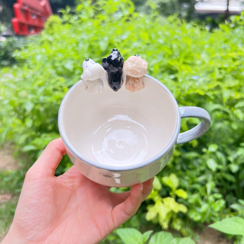 May include: A white ceramic mug with a pale blue handle and rim, decorated with three small dog figurines in white, black, and tan. The mug is held in a hand against a backdrop of green foliage.