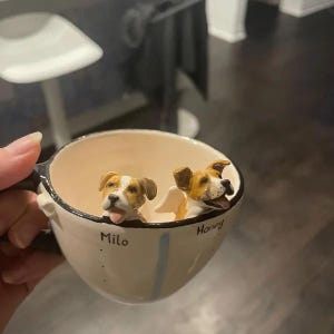May include: A white ceramic mug with a black rim and a blue stripe. Two small dog figurines are inside the mug, one with the name "Milo" and the other with the name "Honey".