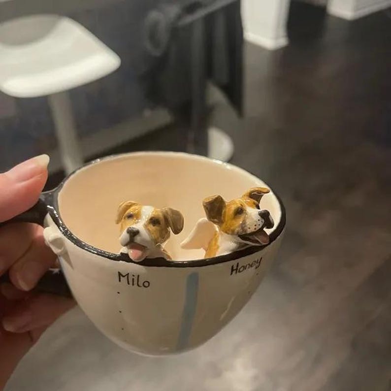 May include: A white ceramic mug with a black rim and a blue stripe. Two small dog figurines are inside the mug, one with the name "Milo" and the other with the name "Honey".