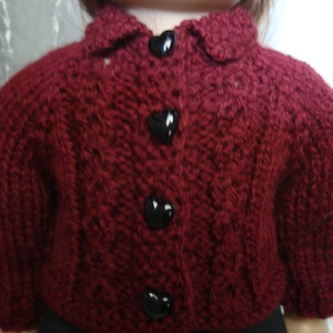 May include: A burgundy knitted cardigan with a collared neckline and three heart-shaped black buttons. The garment features a textured knit pattern and short sleeves, suitable for a doll or small child.