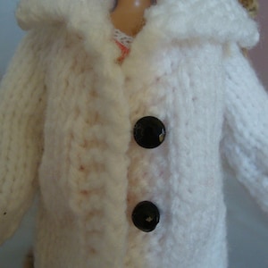 May include: A white hand-knitted coat featuring a wide collar and two round, glossy black buttons down the front. The garment is crafted with a thick, textured yarn stitch pattern and is worn over a light-colored top with a visible lace trim.