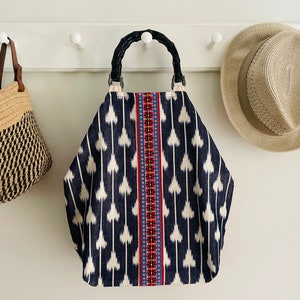 May include: A black and white patterned tote bag with a bamboo handle and colorful embroidered trim. The bag is hanging on a white wall with hooks.