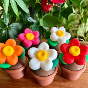 May include: A selection of vibrant, handmade flower decorations. Each flower features a yellow centre, petals in orange, pink, white, and red, and green leaves. They are presented in small, brown terracotta pots, creating a bright, decorative display.
