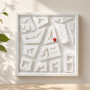 Puede incluir: Obra de arte enmarcada en blanco con un relieve 3D de formas de edificios abstractas. Un pequeño corazón rojo está colocado en una de las estructuras. La obra es de diseño moderno y minimalista.