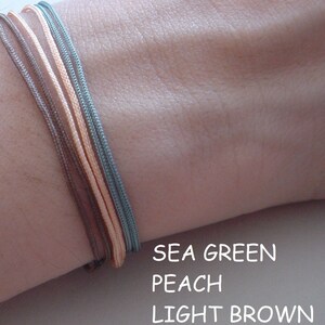 May include: Three bracelets in sea green, peach, and light brown colours. The bracelets are made of cord and are stacked on a person's wrist.