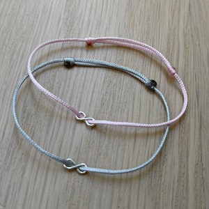 Puede incluir: Dos pulseras de la amistad, una rosa y otra gris, con dijes de símbolo de infinito. Las pulseras están hechas de cordón delgado y están atadas juntas.