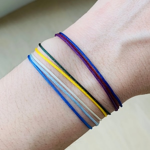 May include: A close-up of a wrist adorned with six thin, colorful bracelets. The bracelets are in shades of blue, light blue, white, yellow, green, and a combination of red and blue. The bracelets are made of a thin, cord-like material.