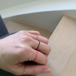 Może przedstawiać: Ręka z trzema cienkimi pierścionkami na palcu serdecznym. Pierścionki są czarne, r&oacute;żowe i złote.