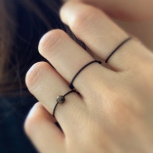 Może przedstawiać: Ręka z trzema cienkimi czarnymi pierścionkami na środkowym i serdecznym palcu. Palec serdeczny ma r&oacute;wnież mały brązowy pierścionek z brązowym kamieniem.