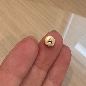 Peut inclure: Une petite perle ronde en métal doré avec la lettre majuscule A gravée en noir sur sa face avant. La perle présente une finition lisse et polie ainsi qu'une forme circulaire, conçue comme un composant pour la création de bijoux ou de loisirs.