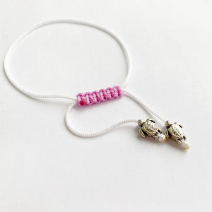 Pode incluir: Pulseira de corda branca com uma seção trançada rosa e dois pingentes de tartaruga prateados.