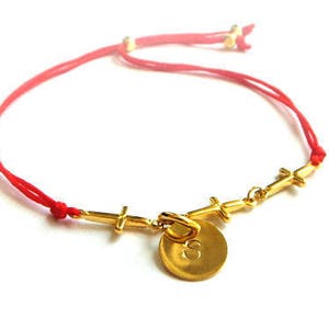 May include: A red string bracelet with a gold charm featuring a circle with the number 8 and four small crosses.