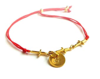May include: A red string bracelet with a gold charm featuring a circle with the number 8 and four small crosses.