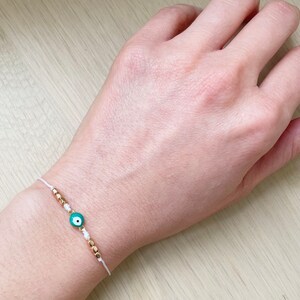 Könnte beinhalten: Weißes Armband aus Schnur mit goldenen Perlen und einem grünen Glücksbringer-Auge.