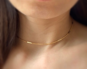 May include: Delicate gold-colored necklace with a fine chain and a small, rectangular bar design. The necklace is worn around the neck, showcasing its minimalist and elegant style. The jewelry is a choker style.