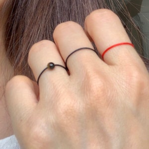 Może przedstawiać: Trzy pierścionki na ręce osoby. Pierścionek na palcu wskazującym to cienki czerwony sznurek. Pierścionek na środkowym palcu to cienki czarny sznurek. Pierścionek na palcu serdecznym to cienki czarny sznurek z małą brązową koralikiem.