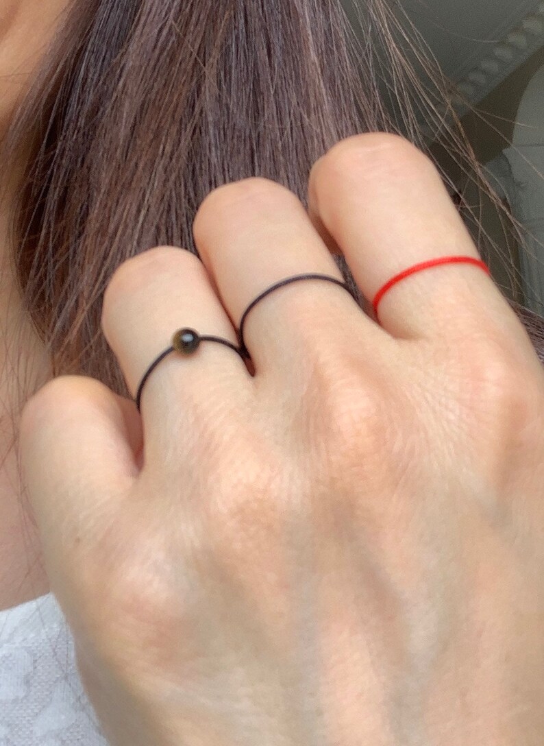 Może przedstawiać: Trzy pierścionki na ręce osoby. Pierścionek na palcu wskazującym to cienki czerwony sznurek. Pierścionek na środkowym palcu to cienki czarny sznurek. Pierścionek na palcu serdecznym to cienki czarny sznurek z małą brązową koralikiem.