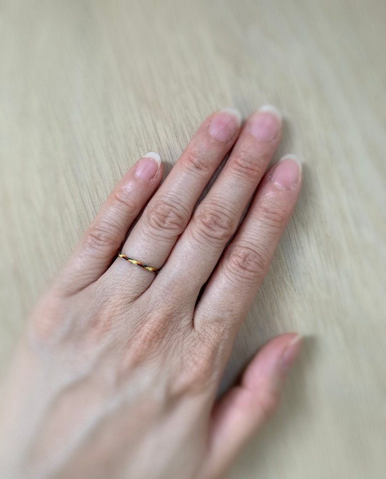 Może przedstawiać: Zbliżenie na dłoń z cienkim, plecionym pierścionkiem z ż&oacute;łtych, pomarańczowych i brązowych nici.