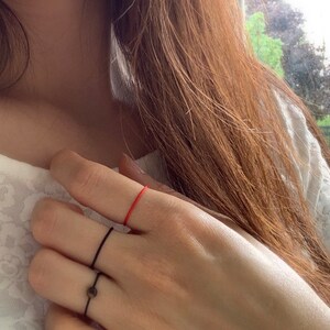 Może przedstawiać: Ręka z trzema pierścionkami na palcu serdecznym. Pierścienie to cienka czarna obręcz, grubsza czarna obręcz z małym okrągłym kamieniem i cienka czerwona sznur&oacute;wka.