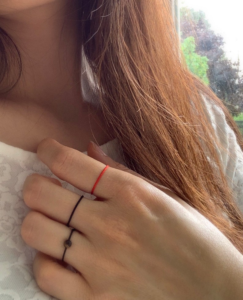 Może przedstawiać: Ręka z trzema pierścionkami na palcu serdecznym. Pierścienie to cienka czarna obręcz, grubsza czarna obręcz z małym okrągłym kamieniem i cienka czerwona sznur&oacute;wka.