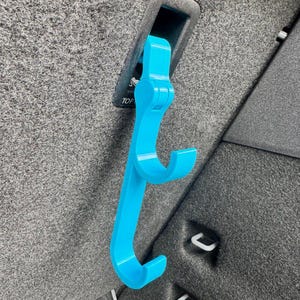 Könnte beinhalten: Ein hellblauer Autositzhaken ist in einen schwarzen rechteckigen Schlitz eingesetzt. Der Haken hat ein gebogenes Design mit zwei unteren Haken. Das Wort "TOP" ist auf dem schwarzen Schlitz sichtbar. Der Haken ist aus Kunststoff.