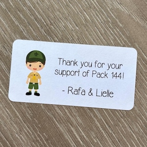May include: White rectangular sticker with rounded corners. The sticker features a cartoon scout with a green hat and uniform. Text on the sticker reads "Thank you for your support of Pack 144! - Rafa & Lielle."