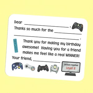 May include: A thank you card with a video game theme. The card has a blank space for the recipient's name and a space to write a message. The card also features a variety of video game controllers and a screen displaying the text "Jackson reached Level 8".