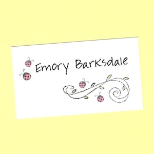 May include: A white card with black text that reads "Emory Barksdale". The card is decorated with a black vine with pink ladybugs.