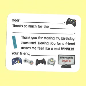 May include: A white card with a handwritten thank you note. The note says "Dear, Thanks so much for the _____, Thank you for making my birthday awesome! Having you for a friend makes me feel like a real WINNER! Your friend, _____." The card is decorated with images of video game controllers and a screen that says "You reached Level 8".