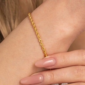 May include: A gold rope chain bracelet is worn on a wrist. The bracelet is a bright, shiny gold color. The bracelet is made of twisted strands of metal. The skin tone is fair with a light pink nail polish.