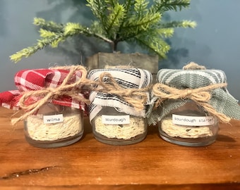 May include: Three small glass jars filled with a light-colored substance, each labeled with a different name. The jars are covered with fabric lids tied with twine. One lid is red and white checkered, one is black and white striped, and the third is green plaid.