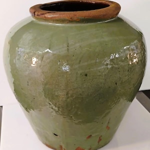 Puede incluir: Un jarrón de cerámica verde de estilo antiguo, con un cuerpo ancho y un borde ligeramente ensanchado. El jarrón tiene un interior marrón oscuro y un borde marrón claro. La superficie muestra signos de edad y desgaste, lo que aumenta su atractivo vintage. Un recipiente de almacenamiento decorativo.