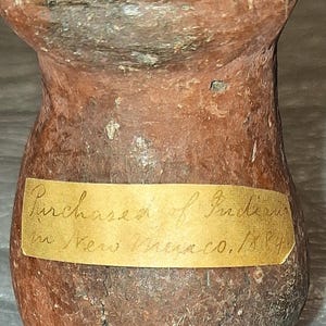 May include: A small, antique, reddish-brown pottery vessel with a rounded base and flared rim. A label on the vessel reads "Purchased of Indians in New Mexico, 1884."