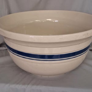 May include: Cream-colored ceramic mixing bowl with a wide rim and two horizontal blue stripes. The bowl has a rounded shape and is designed for mixing ingredients. A classic kitchenware item.