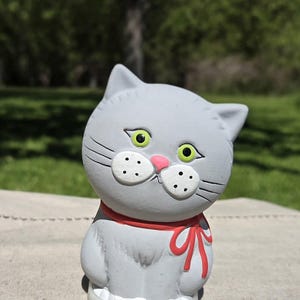 Puede incluir: Una figura de gato de cerámica gris con ojos verde brillante, una pequeña nariz rosa y manchas blancas en el hocico con puntos negros. El gato lleva una cinta roja pintada alrededor del cuello y se asienta sobre una base circular translúcida.